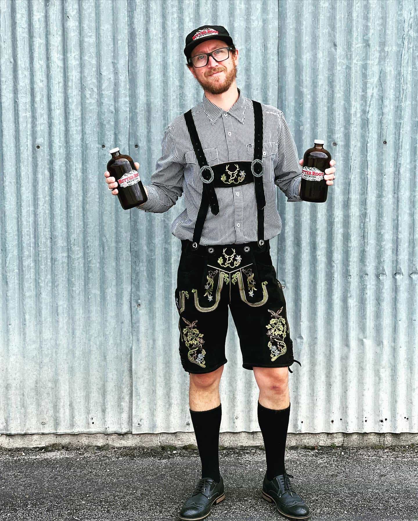 Man in traditional Bavarian attire holding two bottles against a corrugated metal background