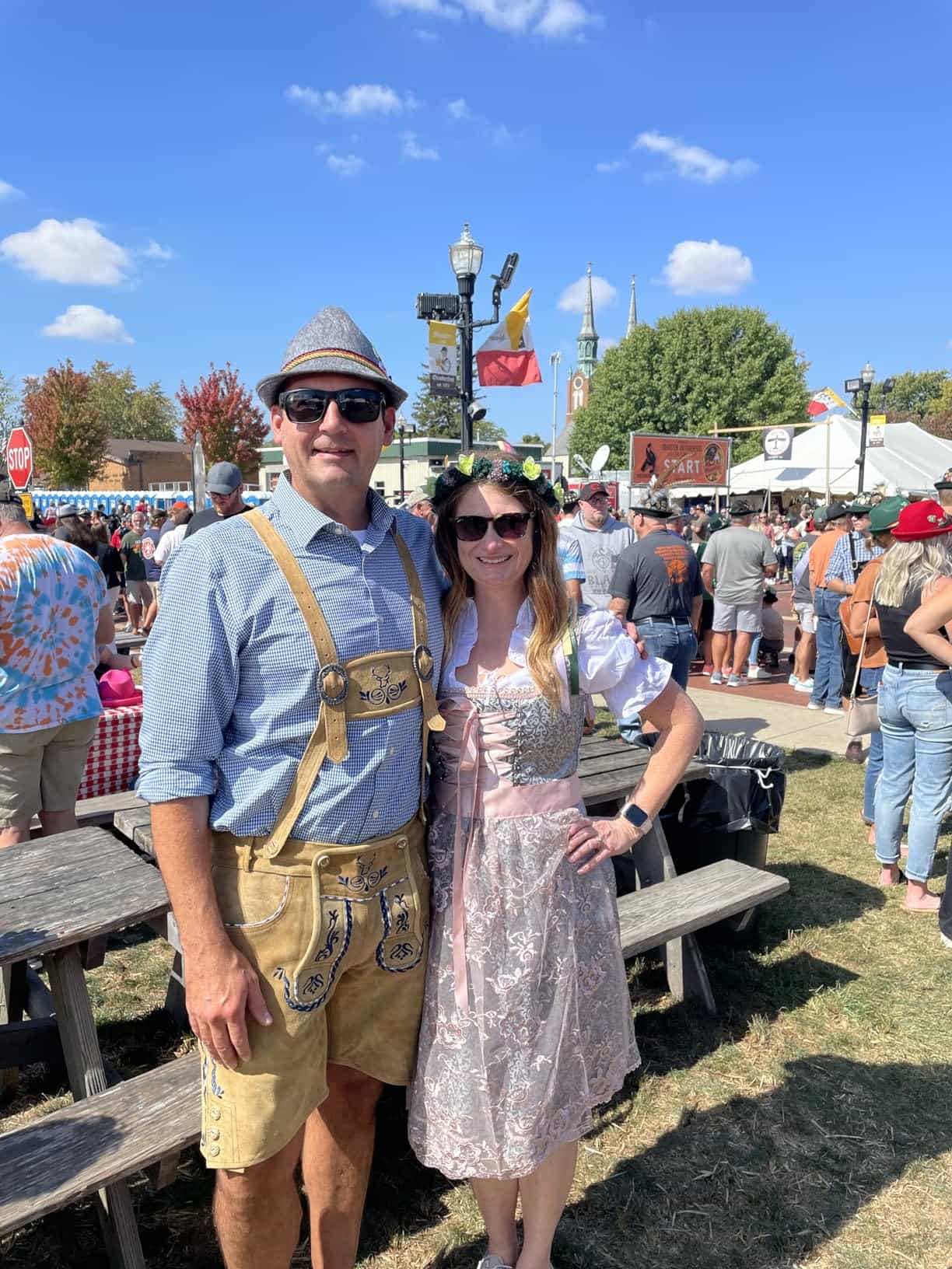 Two people posing together at an outdoor event with a crowd and tents in the background.