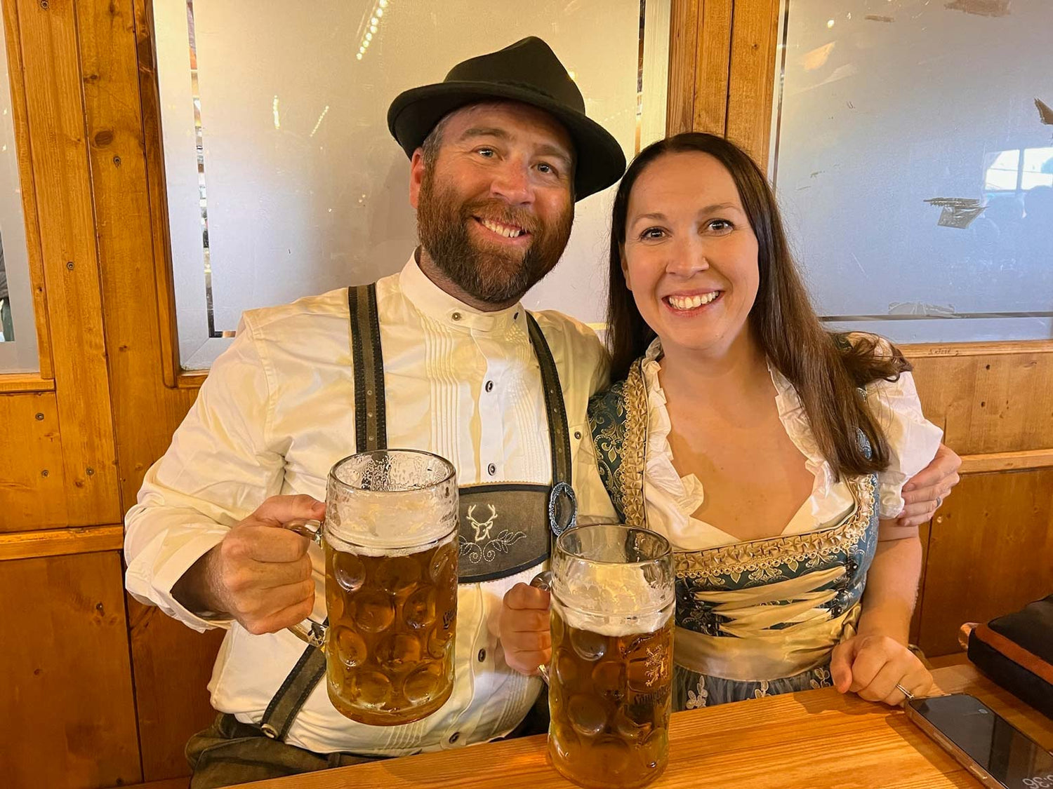 Two people in traditional German clothing with beer mugs in a wooden interior setting.