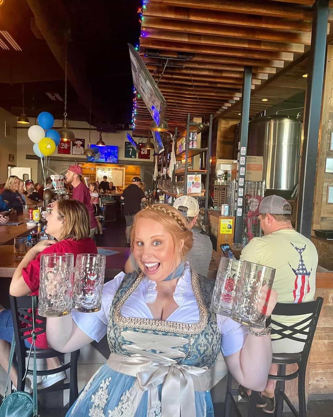 Woman in a traditional Bavarian outfit holding two large steins in a pub setting.