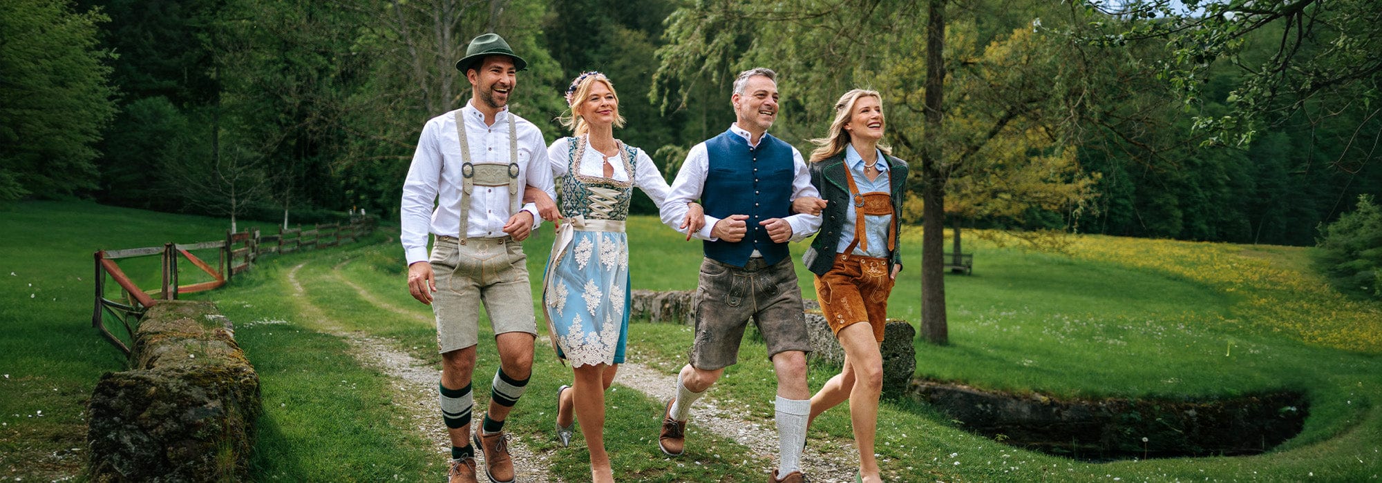 Four people in traditional Bavarian clothing walking on a path in a forest.